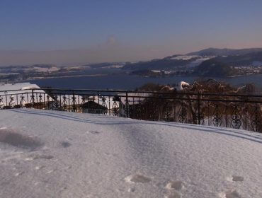 Schneebedeckte Terrasse mit kunstvollem Geländer und Blick auf einen See und ferne Hügel bei klarem Himmel.