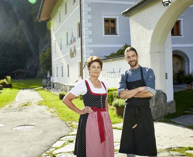 Eine Frau in traditioneller Kleidung und ein Koch stehen vor einem weißen Gebäude, das Essen in Salzburg hervorhebt.