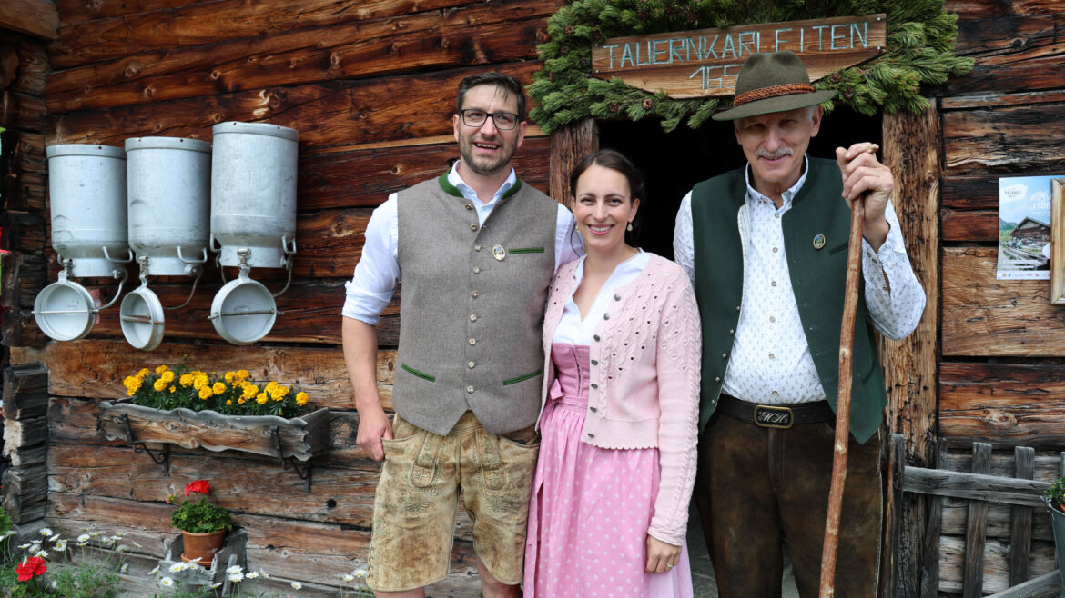 Drei Personen in traditioneller österreichischer Tracht lächeln bei einer Almsommer-Eröffnung vor einem rustikalen Holzgebäude mit Blumen. (vergrößerte Ansicht)