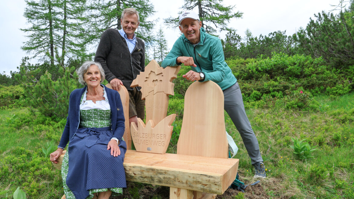 Drei lächelnde Menschen auf und bei Salzburger Almenweg Jubiläumsbank aus Holz während der Almsommereröffnung. Im Hintergrund Bäume (vergrößerte Ansicht)