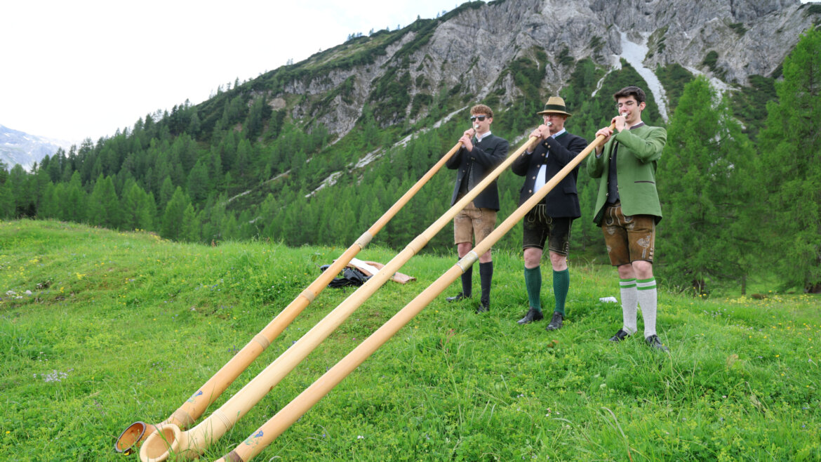 Drei Menschen in traditioneller Tracht, die bei der Almsommer-Eröffnung in einer grasbewachsenen Berglandschaft Alphorn spielen. (vergrößerte Ansicht)