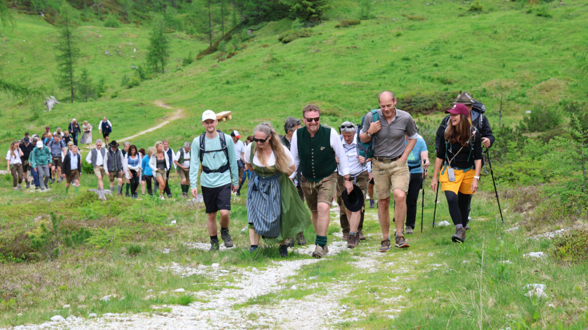 Eine große Gruppe von Menschen wandert anlässlich der Almsommer-Eröffnung auf einem grasbewachsenen, felsigen Pfad inmitten grüner Bäume und Hügel bergauf. (vergrößerte Ansicht)