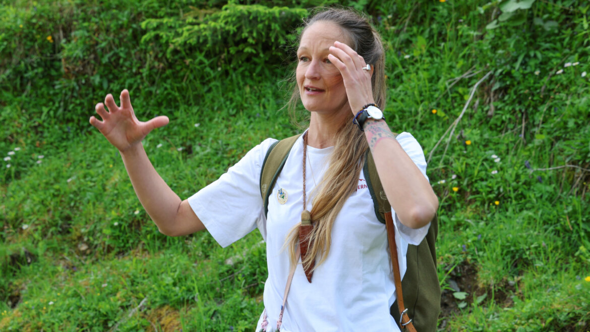 Eine Frau mit langen Haaren in einem weißen T-Shirt gestikuliert bei der Almsommer-Eröffnung in einer grünen, grasbewachsenen Umgebung im Freien. (vergrößerte Ansicht)