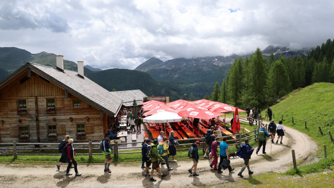 Wanderer gehen während der Almsommer-Eröffnung an einer Almhütte vorbei, mit roten Sonnenschirmen inmitten von Bergen und Bäumen. (vergrößerte Ansicht)