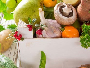 Kiste mit verschiedenen frischen Gemüsesorten und Kräutern wie Pilzen, Knoblauch, Paprika und Kartoffeln auf einem weißen Tisch.