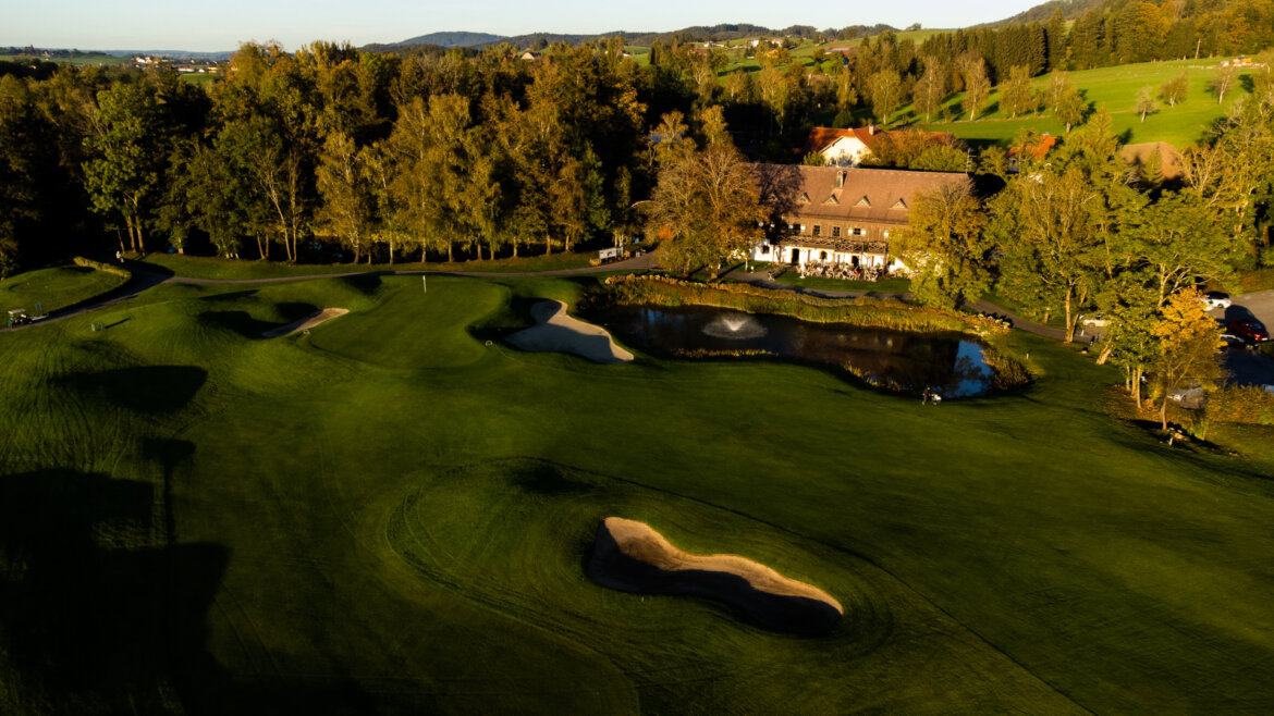 Luftaufnahme eines Golfplatzes mit Sandbunkern, Teich, Clubhaus und umliegenden Bäumen im Sonnenlicht. (vergrößerte Ansicht)