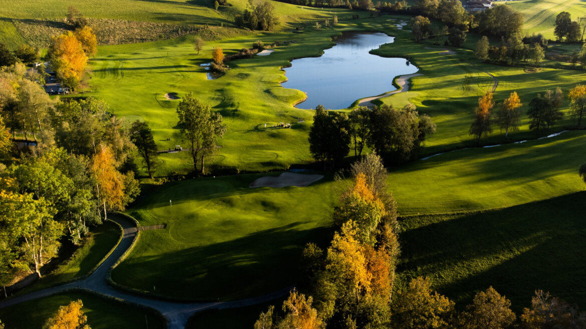 Luftaufnahme eines grünen Golfplatzes mit Bäumen, Sandfängen und einem großen Teich im Sonnenlicht. (vergrößerte Ansicht)