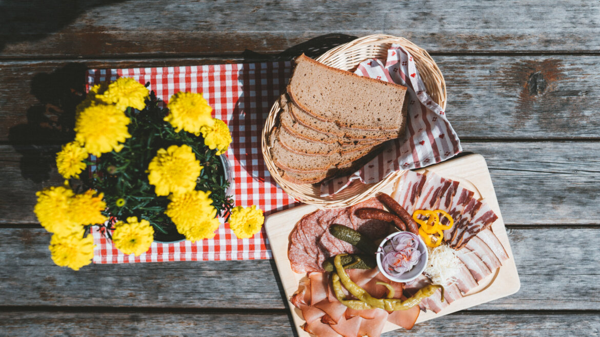 Holztisch mit aufgeschnittenem Brot, Aufschnitt und Essiggurken für die Almsommer-Eröffnung auf einem rot-karierten Tuch. (vergrößerte Ansicht)