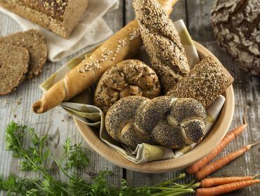 Eine Schale mit verschiedenen Brotsorten und Karotten auf einem rustikalen Holztisch, im Hintergrund weitere Brote und Brotscheiben.