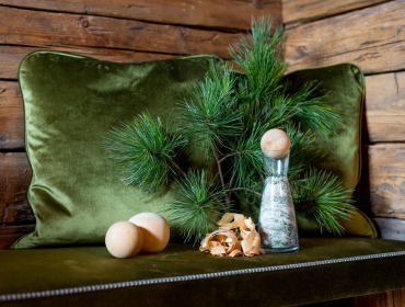 Grünes Kissen mit Kiefernzweigen, einer Flasche mit Kräutern, Holzspänen und Holzkugeln auf einer Bank an einer Holzwand.