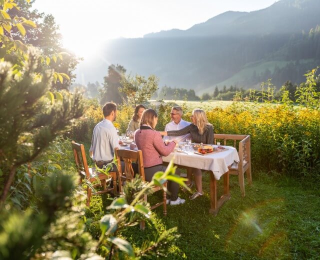 Fünf Personen essen im Freien an einem Tisch in einem üppigen grünen Garten mit Bergen im Hintergrund bei Sonnenuntergang.
