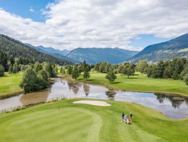 Golfer stehen auf einem Grün in der Nähe eines Sandbunkers mit einem Teich, Bäumen und Bergen im Hintergrund unter einem bewölkten Himmel.