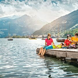 Eine Familie entspannt sich auf einem Holzsteg am See, umgeben von Segelbooten und Bergen, während ein Mann einem Kind den Fahrradhelm richtet.