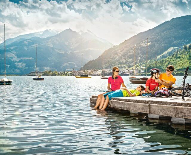Tauernradweg Pause am See, Markus Greber Eine Familie entspannt sich auf einem Holzsteg am See, umgeben von Segelbooten und Bergen, während ein Mann einem Kind den Fahrradhelm richtet.