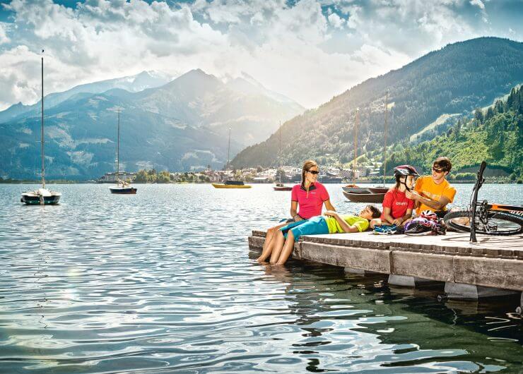 Eine Familie entspannt sich auf einem Holzsteg am See, umgeben von Segelbooten und Bergen, während ein Mann einem Kind den Fahrradhelm richtet.