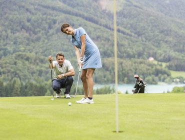 Frau beim Putten auf einem Golfplatz mit einem Mann, der hinter ihr hockt; Berge und See im Hintergrund.