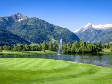 Zell_am_See_-15 Golfplatz mit Wasserfontäne, See, grünen Bäumen und Bergen unter einem strahlend blauen Himmel.