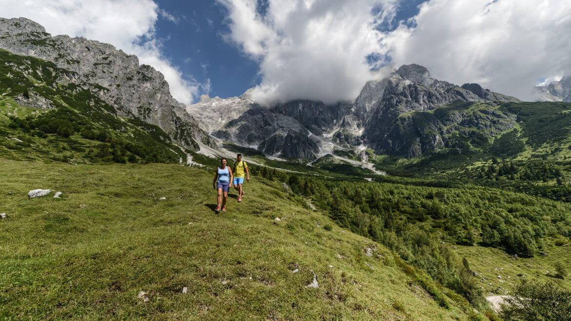 Zwei Wanderer gehen auf einem grasbewachsenen Hügel mit Bergen und einem teilweise bewölkten Himmel im Hintergrund.