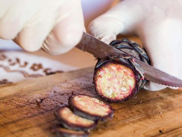 Mit Handschuhen behandelte Hände, die mit einem gezackten Messer eine violette Kaktusfrucht auf einem hölzernen Schneidebrett schneiden.
