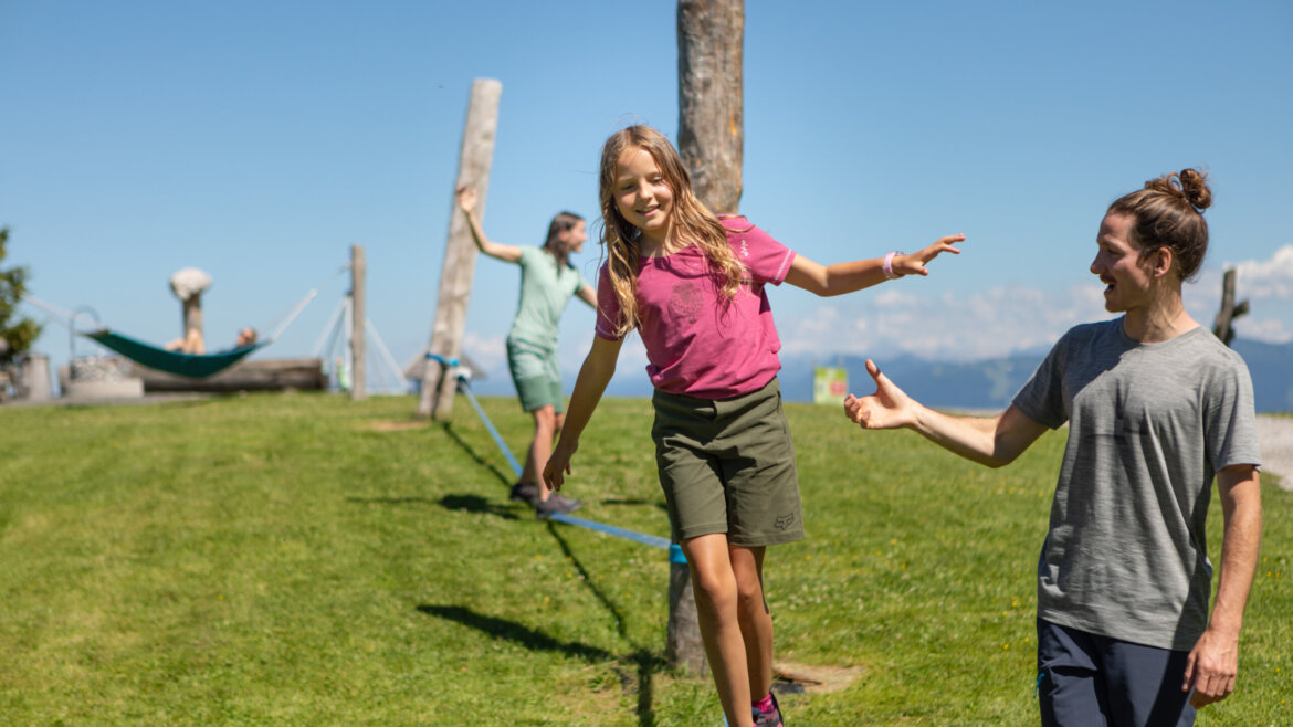 Ein Mädchen balanciert draußen auf einer Slackline, während ein Mann ihr Halt gibt und eine weitere Person im Hintergrund zu sehen ist. (vergrößerte Ansicht)