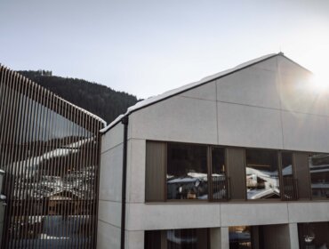 Die Sonne scheint über moderne Gebäude mit großen Fenstern, Schnee auf den Dächern und Bergen im Hintergrund.