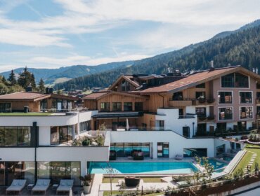 Ein modernes Bergresort mit Außenpool, umgeben von Bäumen und Bergen unter blauem Himmel.