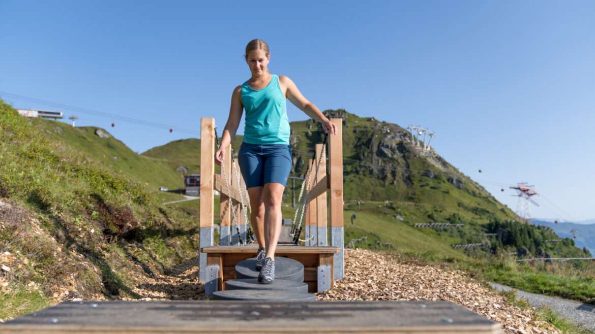 © Gasteiner Bergbahnen AG Christoph Oberschneider Eine Frau balanciert auf einer Holzbrücke im Freien in einer bergigen Gegend unter strahlend blauem Himmel. (vergrößerte Ansicht)