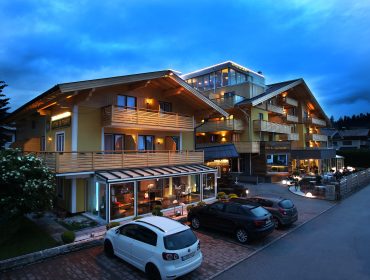 Modernes Hotelgebäude mit warmen Lichtern in der Abenddämmerung, davor geparkte Autos, darüber bewölkter Himmel.