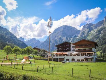 Ein großes Chalet mit Balkonen liegt auf einer grünen Wiese, umgeben von Bergen unter einem blauen Himmel mit Wolken.