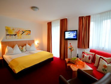 Helles Hotelzimmer mit Doppelbett, rotem Sofa, TV und großen Fenstern mit transparenten Vorhängen.