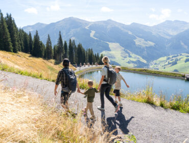 Vierköpfige Familie, die auf einem Bergpfad an einem See spazieren geht, umgeben von Bäumen und fernen Bergen.
