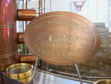 Ovales Madlberggut-Metallschild mit der Aufschrift "Dachstein-Destillerie" und eingravierten Bergen, in einer Brennereiumgebung.