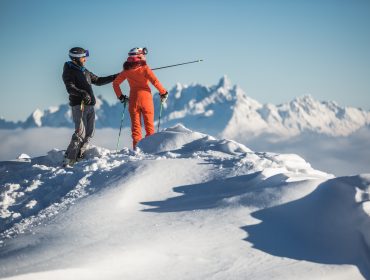Meisis-Reise-Flachau-Hermann-Maier-zeigt-Alexandra-Meissnitzer-den-Ausblick-auf-das-Skigebiet-Flachau Zwei Skifahrer stehen auf einem verschneiten Berggipfel in Flachau und blicken auf ferne schneebedeckte Gipfel unter einem klaren Himmel.
