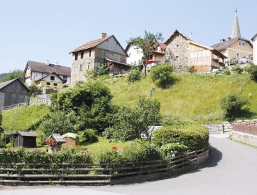 Mitten im Leben – Foto Dreiländerwirt Ein Dorf auf einem Hügel mit Steinhäusern, Grünflächen, Holzzäunen und einem Kirchturm unter einem klaren Himmel.