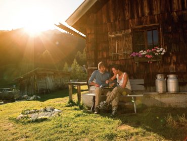 Ein Paar sitzt bei Sonnenuntergang auf einer Bank bei einer rustikalen österreichischen Hütte und genießt ein Getränk in einer österreichischen Berglandschaft.