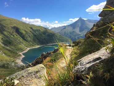 Bergpfad im Nassfeld mit Blick auf einen blauen Alpensee, grüne Hügel und einen fernen Gipfel unter einem strahlenden Himmel.