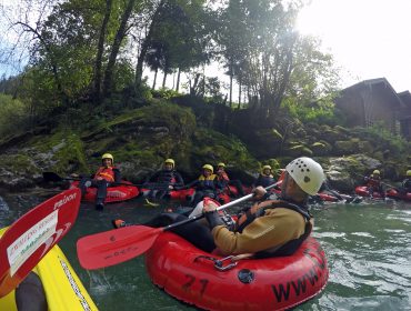 Eine Gruppe mit Helmen und Neoprenanzügen sitzt in roten aufblasbaren Schläuchen auf dem Fluss Lammer, umgeben von Bäumen.
