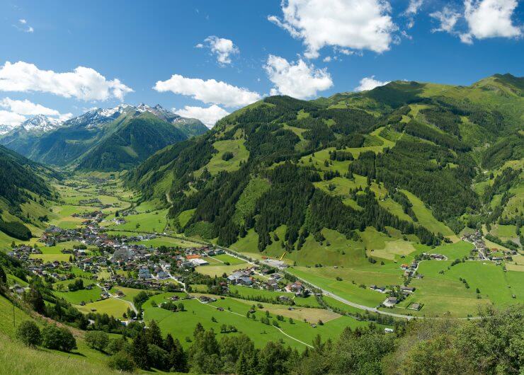 Panoramablick auf ein grünes Tal mit einer kleinen Stadt, umgeben von Bergen unter einem blauen Himmel mit Wolken.