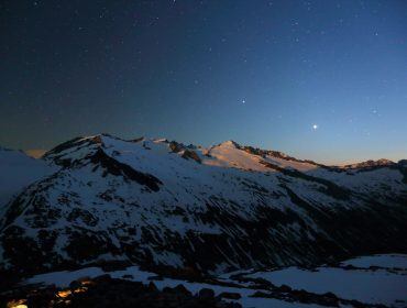 Schneebedeckte Berge unter einem sternenklaren Nachthimmel mit einem hellen Planeten über den Gipfeln.