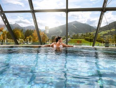 Eine Frau, die sich an einem sonnigen Tag in einem Thermenpool mit Blick auf die Berge durch große Glasfenster entspannt.