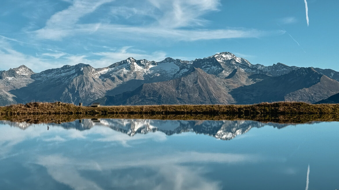 © Gasteiner Bergbahnen AG Schneebedeckte Berge spiegeln sich in einem ruhigen See unter einem blauen Himmel mit vereinzelten Wolken. (vergrößerte Ansicht)