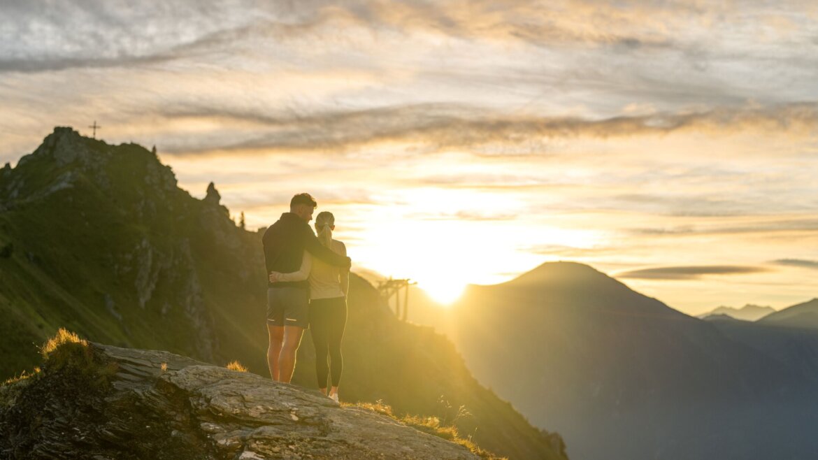 © Gasteiner Bergbahnen AG Zwei Menschen stehen bei Sonnenaufgang auf einem Berg und umarmen sich, während sie den Blick über eine malerische, sonnenbeschienene Landschaft schweifen lassen. (vergrößerte Ansicht)