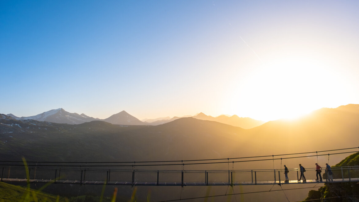 © Gasteiner Bergbahnen AG Menschen überqueren bei Sonnenuntergang eine Hängebrücke vor dem Hintergrund der Berge. (vergrößerte Ansicht)