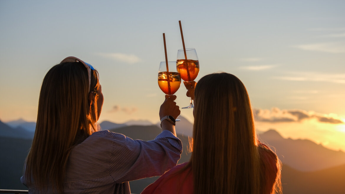 © Gasteiner Bergbahnen AG Zwei Frauen stoßen bei Sonnenuntergang mit einem Drink an und genießen den Blick auf die Berge und den leuchtenden Himmel. (vergrößerte Ansicht)