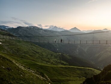 Menschen, die bei Sonnenuntergang über eine lange Hängebrücke über ein grünes Bergtal gehen, im Hintergrund sind Berggipfel zu sehen.