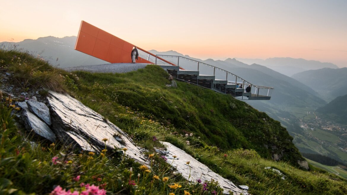 © Gasteiner Bergbahnen AG Marktl Photography Eine Person steht bei Sonnenuntergang auf einem orangefarbenen Aussichtspunkt, im Vordergrund sind Blumen und Berge zu sehen. (vergrößerte Ansicht)