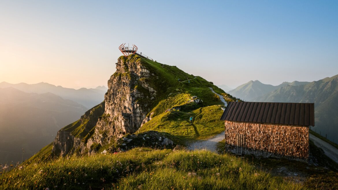 © Gasteiner Bergbahnen AG Marktl Photography Eine Holzhütte und ein grasbewachsener Hügel mit einer Aussichtsplattform auf einem Felsgipfel unter klarem Himmel bei Sonnenuntergang. (vergrößerte Ansicht)