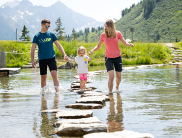 Eine dreiköpfige Familie, die sich an den Händen hält, geht auf Trittsteinen über einen seichten Bach in einer Berglandschaft.