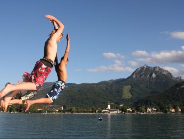 Zwei Jungen in Badehosen springen in den Wolfgangsee mit Bergen und Gebäuden im Hintergrund unter einem blauen Himmel.