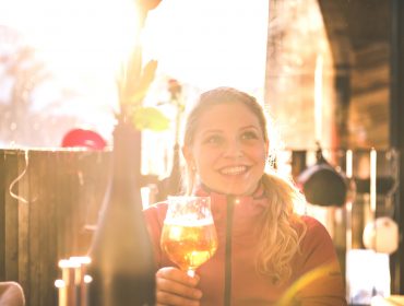 Wintershooting_2018-19_Scha Lächelnde Frau im Sonnenlicht mit einem Glas Bier in einem Straßencafé in Hochkönig.